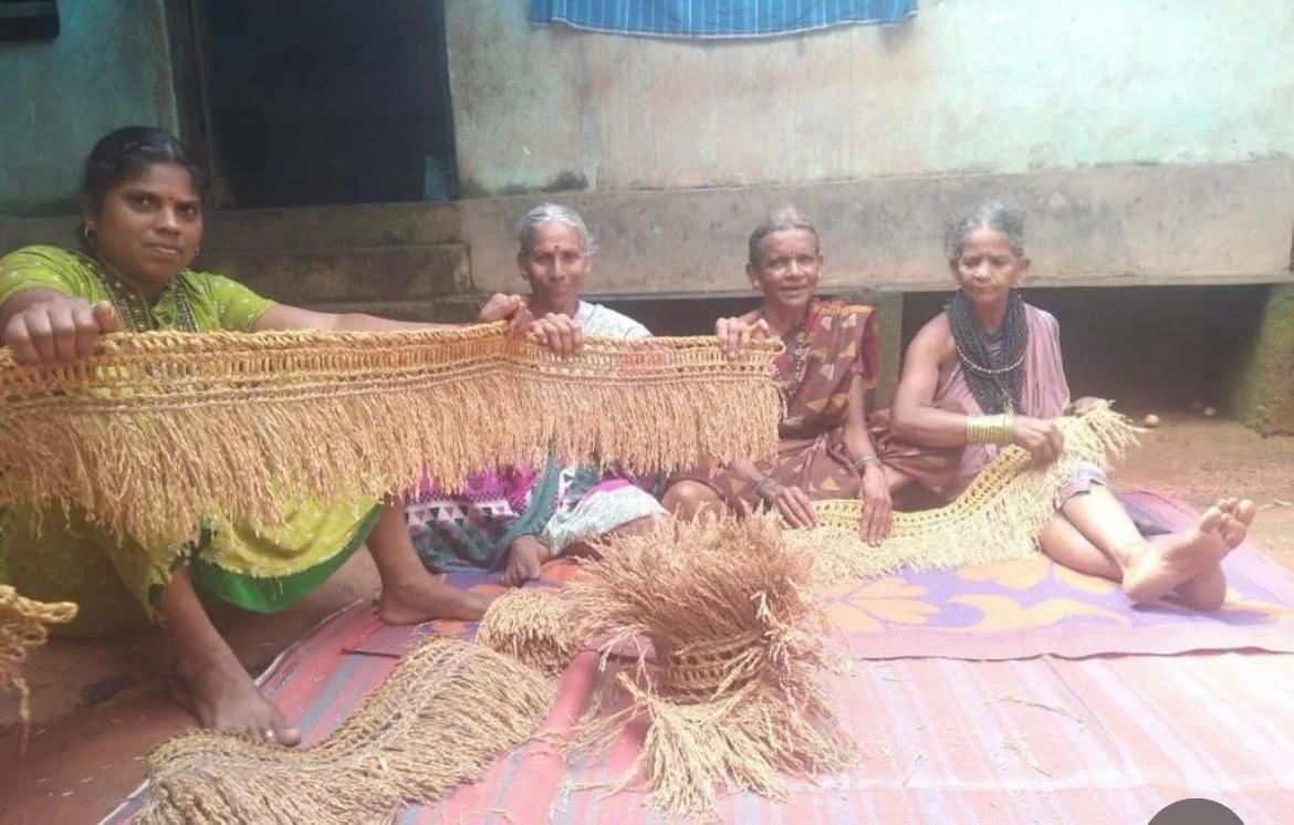Four women artisans sitting on a mat outside a house, holding up long paddy torans
