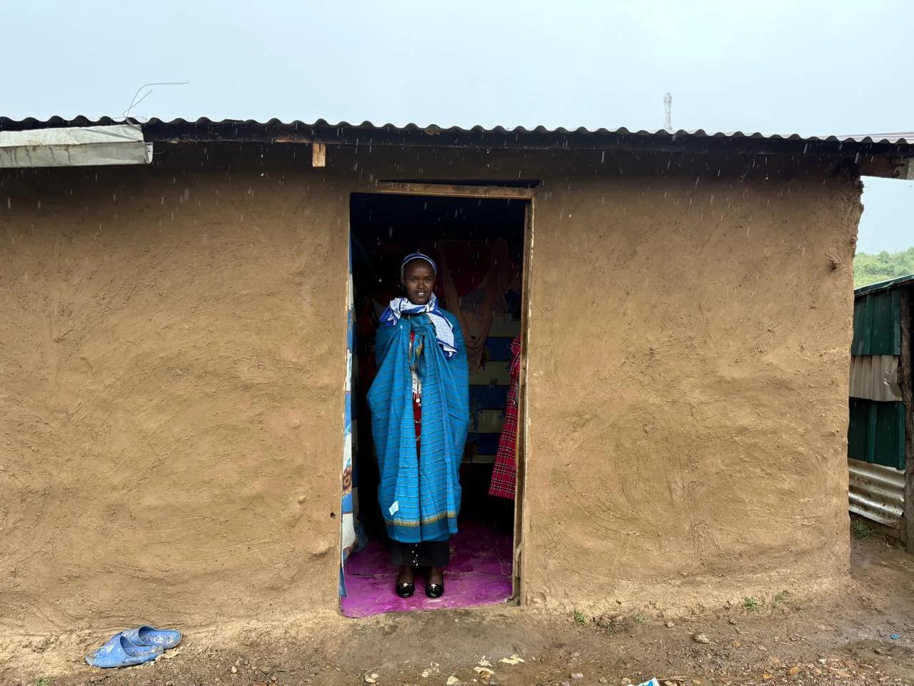 A Maasai woman in a blue shúkà standing in the doorway of a mud house in Talek